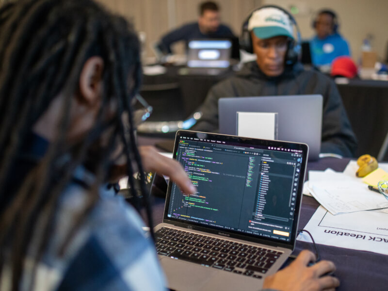 Close-up of a laptop screen displaying code, with a blurred user in the foreground and other hackathon participants working on laptops in the background.